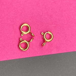 May include: Three gold-toned spring ring clasps for jewelry making. Each circular clasp features a small spring-loaded lever mechanism and a fixed loop attachment point, designed for securing necklaces or bracelets with a reliable closure.