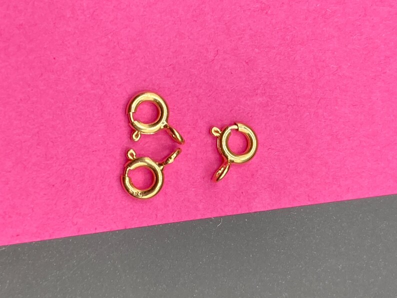 May include: Three gold-toned spring ring clasps for jewelry making. Each circular clasp features a small spring-loaded lever mechanism and a fixed loop attachment point, designed for securing necklaces or bracelets with a reliable closure.