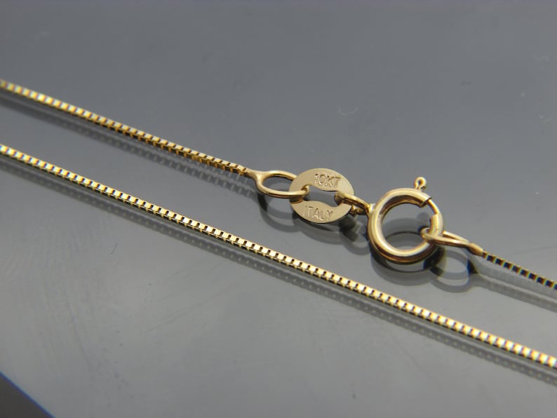 May include: Close-up of a gold-colored box chain necklace with a lobster clasp. The clasp and chain are connected by a small ring. The necklace has a delicate, square-link design. The clasp has the text "10K ITALY".