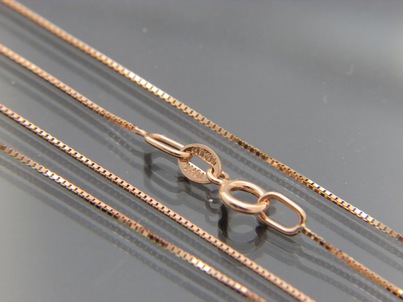 May include: A close-up of a rose gold chain necklace with a lobster clasp. The chain is made of small, square links.