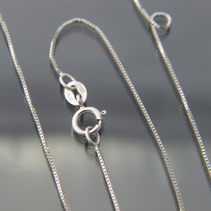 May include: A close-up of a silver box chain necklace with a lobster clasp. The clasp is marked "10KT ITALY". The chain is made of small, square links and is displayed against a dark background.