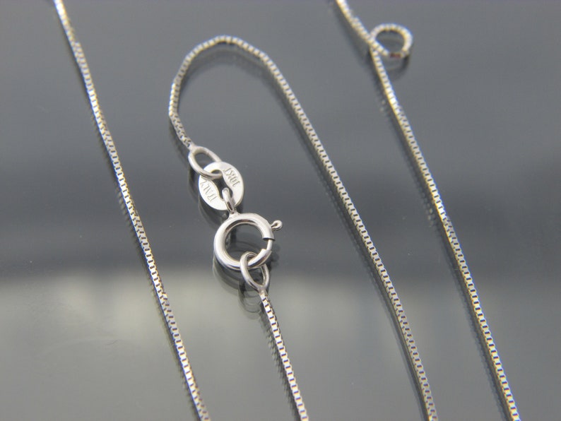 May include: A close-up of a silver box chain necklace with a lobster clasp. The clasp is marked "10KT ITALY". The chain is made of small, square links and is displayed against a dark background.