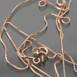 May include: A close-up of a rose gold chain necklace with a lobster clasp. The chain is made of small, square links.