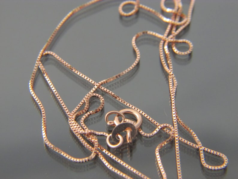 May include: A close-up of a rose gold chain necklace with a lobster clasp. The chain is made of small, square links.
