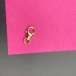May include: A gold-toned metal lobster claw clasp featuring a spring-loaded mechanism and a small circular jump ring attached to the base. The jewelry finding has a polished, reflective surface and is designed for connecting chains or necklaces.