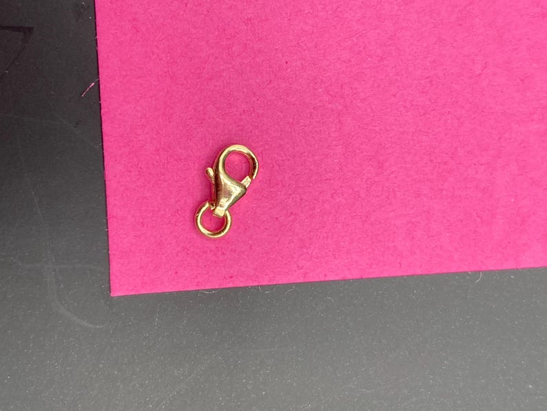 May include: A gold-toned metal lobster claw clasp featuring a spring-loaded mechanism and a small circular jump ring attached to the base. The jewelry finding has a polished, reflective surface and is designed for connecting chains or necklaces.