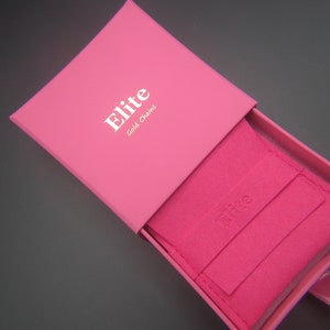 May include: A vibrant pink jewellery box, branded with "Elite Gold Chains" in white. Inside, a pink pouch is visible, also bearing the brand name. A pink ribbon adds a finishing touch.