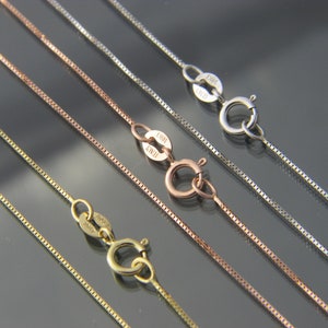 May include: Close-up of several delicate box chain necklaces in gold, rose gold, and silver tones. Each chain features a lobster clasp closure. The chains are displayed against a dark, reflective surface.