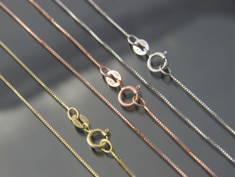 May include: Close-up of several delicate box chain necklaces in gold, rose gold, and silver tones. Each chain features a lobster clasp closure. The chains are displayed against a dark, reflective surface.