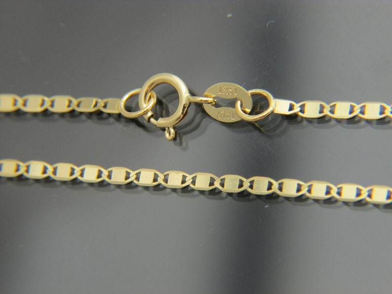 May include: A gold necklace with a flat-link chain design. The necklace has a lobster clasp and a tag marked "14KT ITALY". The chain is made of small, rectangular links. The necklace is likely made of 14-carat gold.