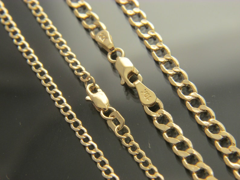 K&ouml;nnte beinhalten: Drei goldene Ketten in verschiedenen Breiten, alle im Curb-Link-Stil. Die Ketten liegen in einer Reihe, wobei die dickste Kette unten und die d&uuml;nnste Kette oben liegt. Die Ketten sind alle aus Gold und haben eine gl&auml;nzende Oberfl&auml;che.