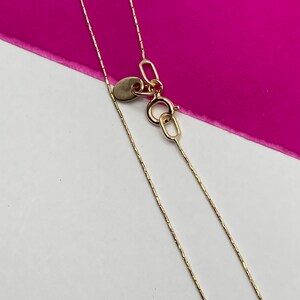 May include: A close-up shot of a fine gold necklace with a thin chain. The necklace has a small, circular tag and a lobster clasp. The background is a bright pink and white.