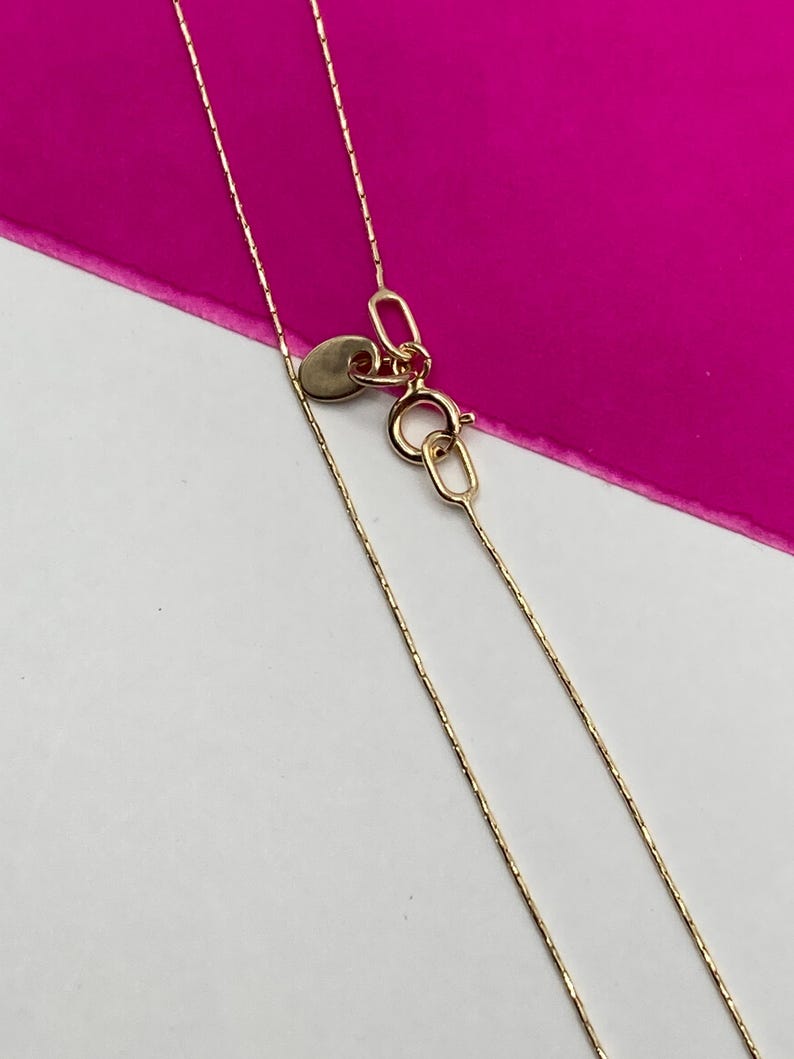 May include: A close-up shot of a fine gold necklace with a thin chain. The necklace has a small, circular tag and a lobster clasp. The background is a bright pink and white.