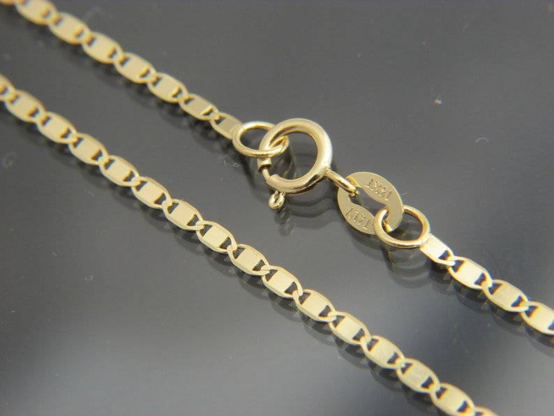 May include: Close-up of a gold necklace with a delicate, flat link chain design. The necklace features a lobster clasp closure and a small tag with the marking "14KT". The chain is approximately 45 cm long.