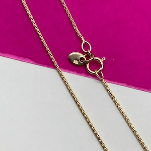 May include: Close-up of a gold chain necklace with a lobster clasp. The chain features a delicate, woven design. The background is a combination of white and bright pink.