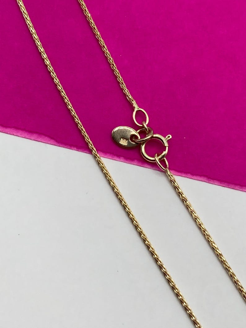 May include: Close-up of a gold chain necklace with a lobster clasp. The chain features a delicate, woven design. The background is a combination of white and bright pink.