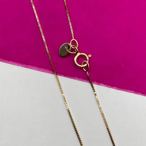 May include: A gold-coloured necklace with a fine chain and a heart-shaped tag. The necklace has a lobster clasp. The background is a bright pink and white.