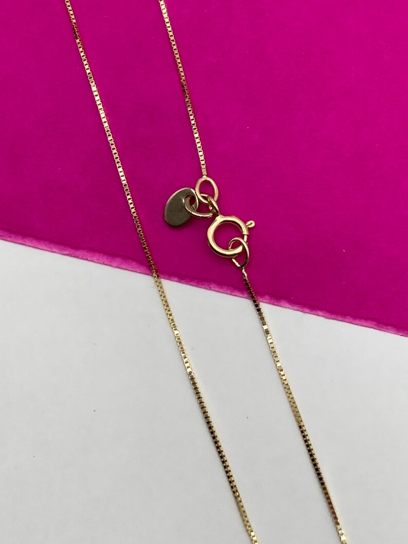 May include: A gold-coloured necklace with a fine chain and a heart-shaped tag. The necklace has a lobster clasp. The background is a bright pink and white.