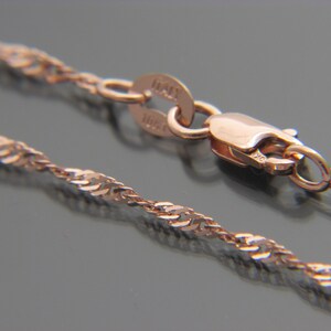 May include: A close-up of a rose gold chain necklace with a twisted rope design. The necklace has a lobster clasp closure and a small tag that reads "ITALY". The chain is displayed on a dark surface.