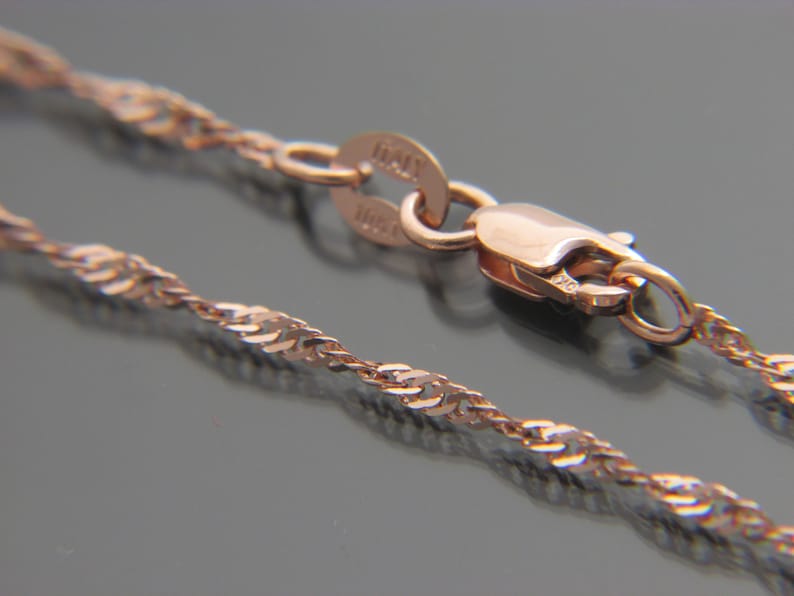 May include: A close-up of a rose gold chain necklace with a twisted rope design. The necklace has a lobster clasp closure and a small tag that reads "ITALY". The chain is displayed on a dark surface.