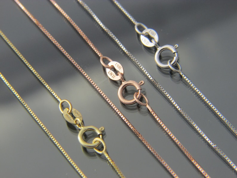 May include: Close-up of several delicate box chain necklaces in gold, rose gold, and silver tones. Each chain features a small clasp and is displayed against a dark, reflective surface. The chains are of varying lengths and thicknesses.