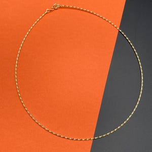 Puede incluir: Un collar de cadena fino de color dorado que presenta una textura delicada de cuerda trenzada a lo largo de toda su longitud. El collar se asegura con un pequeño cierre de resorte circular situado en la parte superior del círculo.