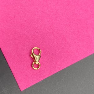 May include: A small, polished gold-tone lobster claw clasp featuring a spring-loaded mechanism and a circular jump ring attached at the base. The metal component has a smooth, reflective surface and a teardrop-shaped body design for jewelry.