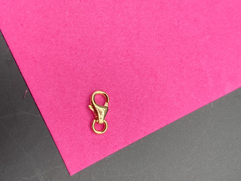 May include: A small, polished gold-tone lobster claw clasp featuring a spring-loaded mechanism and a circular jump ring attached at the base. The metal component has a smooth, reflective surface and a teardrop-shaped body design for jewelry.
