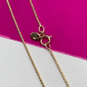 May include: A gold-coloured necklace with a delicate chain design. The necklace features a lobster clasp closure and a small, round tag near the clasp. The background is a vibrant pink and white.
