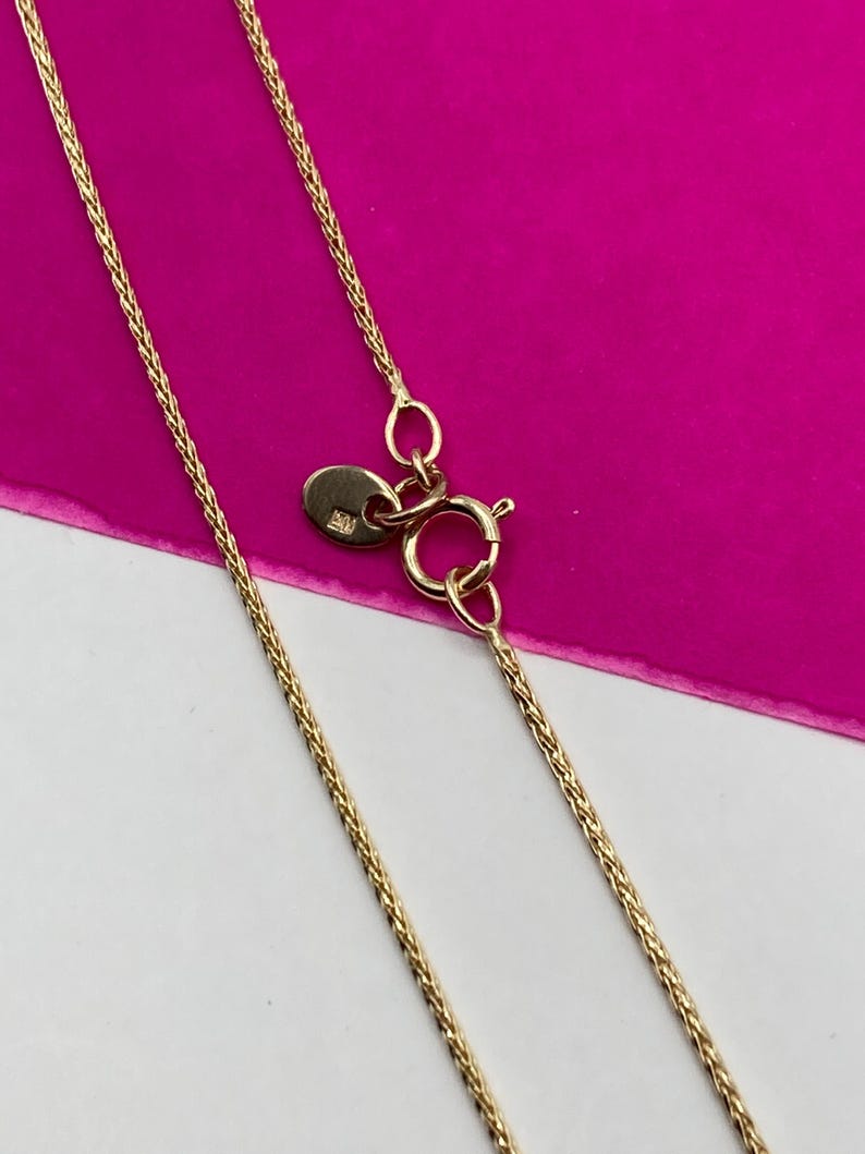 May include: A gold-coloured necklace with a delicate chain design. The necklace features a lobster clasp closure and a small, round tag near the clasp. The background is a vibrant pink and white.