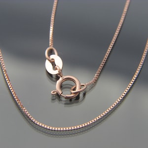 May include: Rose gold box chain necklace with a lobster clasp. The chain is made of small, square links and is displayed against a dark, reflective surface. The clasp is open.