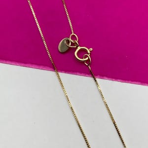 May include: A fine gold necklace with a box chain design. The necklace has a small heart-shaped tag and a lobster clasp. The background is a vibrant pink and white.