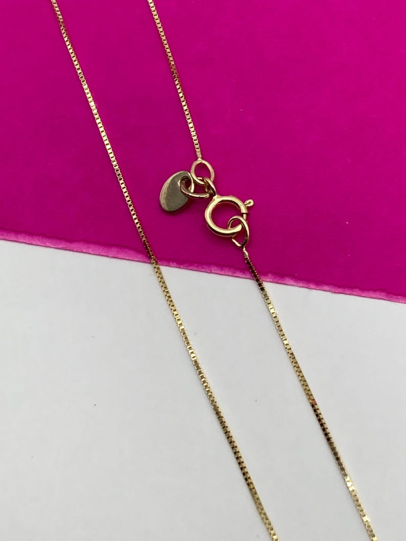 May include: A fine gold necklace with a box chain design. The necklace has a small heart-shaped tag and a lobster clasp. The background is a vibrant pink and white.