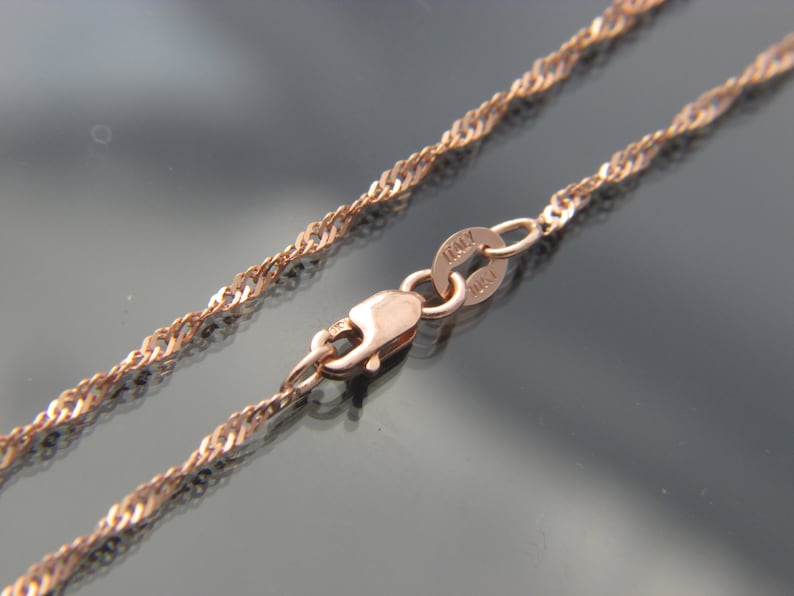 May include: Rose gold-colored necklace with a twisted rope chain design. The clasp and connecting ring are visible, with the word "ITALY" stamped on the ring. The chain is displayed against a dark, reflective surface.