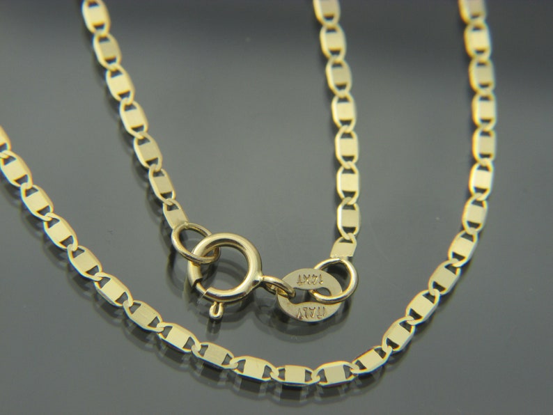 May include: A close-up of a gold necklace with a delicate chain design. The chain features a series of rectangular links. The clasp is visible, and the necklace appears to be made of 14K gold. The necklace is a piece of jewellery.