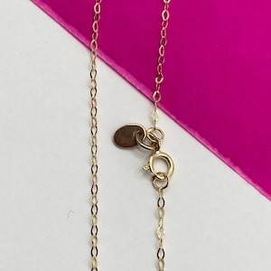 May include: Close-up of a delicate gold necklace with a small oval tag and a spring ring clasp. The chain is made of small, interconnected oval links. The background is a combination of white and bright pink.