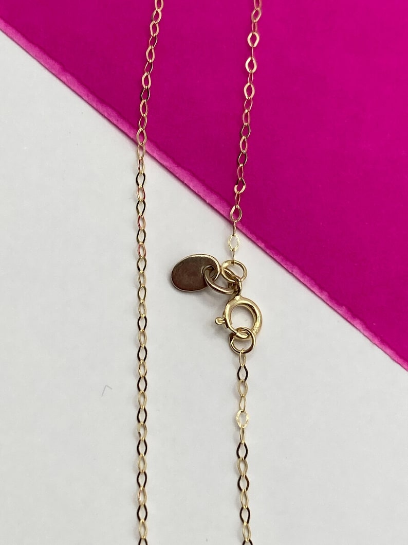 May include: Close-up of a delicate gold necklace with a small oval tag and a spring ring clasp. The chain is made of small, interconnected oval links. The background is a combination of white and bright pink.