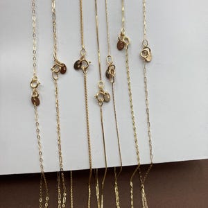 May include: A selection of fine gold necklaces presented against a white backdrop. The image showcases different chain designs, such as box, cable, and rope. Each necklace has a clasp and a small, circular tag. The chains vary in length and thickness.