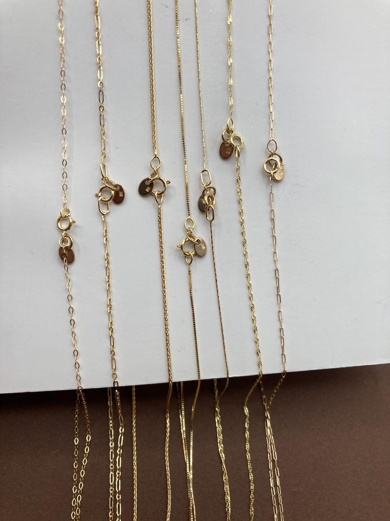 May include: A selection of fine gold necklaces presented against a white backdrop. The image showcases different chain designs, such as box, cable, and rope. Each necklace has a clasp and a small, circular tag. The chains vary in length and thickness.