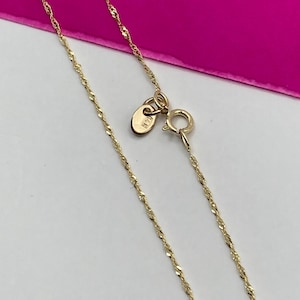 May include: A delicate gold-coloured necklace with a twisted rope chain design. The necklace features a small clasp and a tag near the clasp. The background is a bright pink colour.