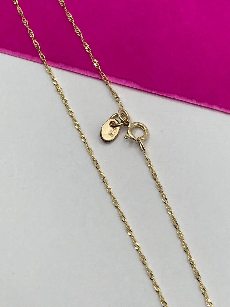 May include: A delicate gold-coloured necklace with a twisted rope chain design. The necklace features a small clasp and a tag near the clasp. The background is a bright pink colour.