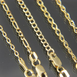Puede incluir: Cinco cadenas de oro de diferentes estilos y grosores. Las cadenas están dispuestas en fila sobre una superficie negra.