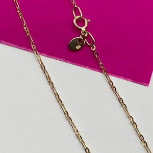 May include: A gold-coloured necklace with a delicate chain design. The necklace features a lobster clasp closure and a small tag near the clasp. The chain has a repeating pattern of elongated and oval links.