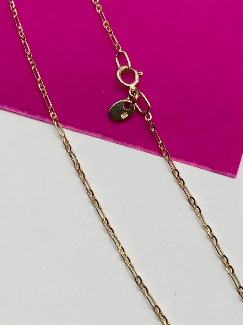 May include: A gold-coloured necklace with a delicate chain design. The necklace features a lobster clasp closure and a small tag near the clasp. The chain has a repeating pattern of elongated and oval links.