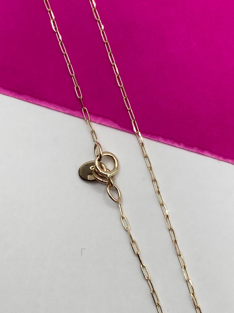 May include: Close-up of a delicate gold necklace with a small, round clasp and a chain of elongated links. The necklace is set against a bright pink background, creating a contrast that highlights the jewelry's details.