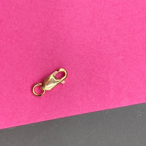 May include: A gold-toned lobster claw clasp featuring a smooth, polished finish and a small circular jump ring attached to one end. The mechanism includes a spring-loaded lever that opens and closes to secure jewelry pieces like necklaces or bracelets.