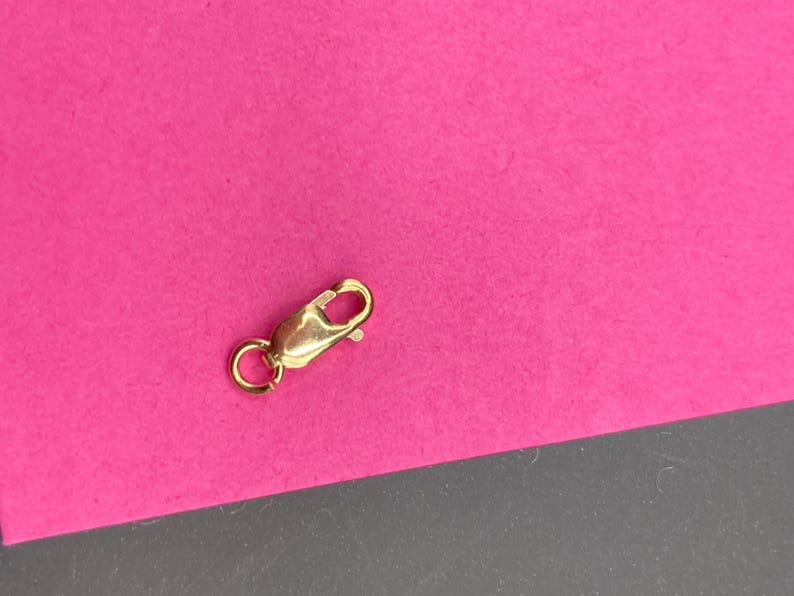 May include: A gold-toned lobster claw clasp featuring a smooth, polished finish and a small circular jump ring attached to one end. The mechanism includes a spring-loaded lever that opens and closes to secure jewelry pieces like necklaces or bracelets.