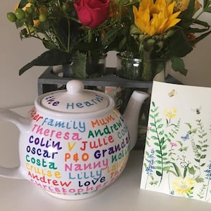 May include: A white teapot with a colourful design featuring the names of family members. The teapot is decorated with the words "The Heart" and "Family Mum Theresa Andrew Colin V Julie Costa Oscar P&O Granda Nanny Holiday Cruise Lilly Love Andrew Christopher".