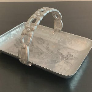 May include: A rectangular silver-toned metal basket with a scalloped edge and a single arched handle. The handle features a repeating floral motif, and the base of the basket is embossed with a central floral design featuring tulips and leaves.