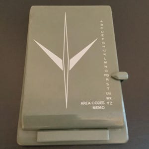 May include: A rectangular olive green plastic memo device featuring a white stylized arrow graphic on the left and a vertical alphabet index from A to Z on the right. The text AREA CODES MEMO is printed in white near the bottom right corner.